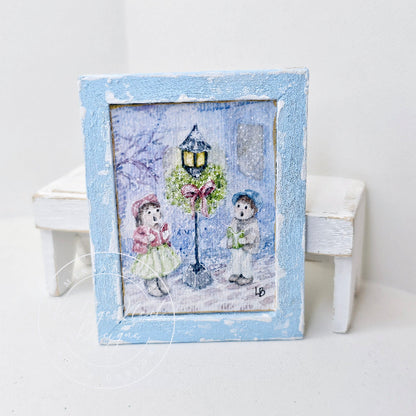 A small square framed art piece with a distressed light blue frame, featuring a whimsical winter scene of two children under a decorated streetlamp. The frame is displayed on a white surface with miniature white stools in the background.